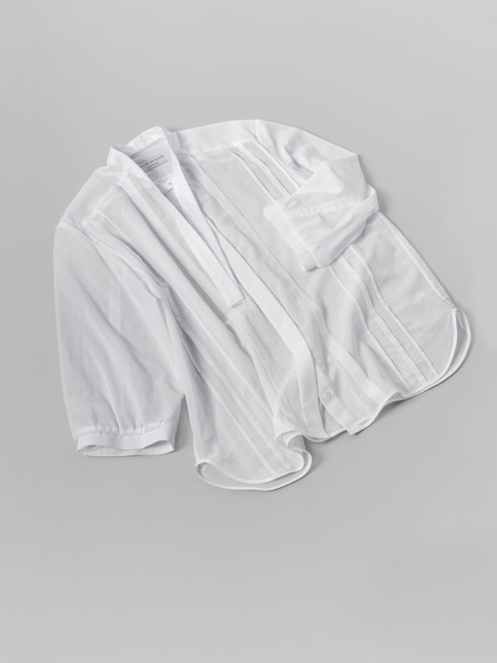 une chemise est posée seule sur le sol. La couleur du sol est à peine plus sombre que la chemise, mais l'on peut voir les détails du vêtement. La chemise est en voile blanc et les manches sont bouffantes.