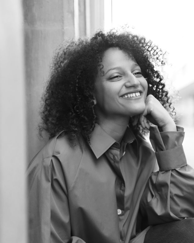 une photo en noir et blanc, où l'on voit une femme souriante , accoudée sur son genou.